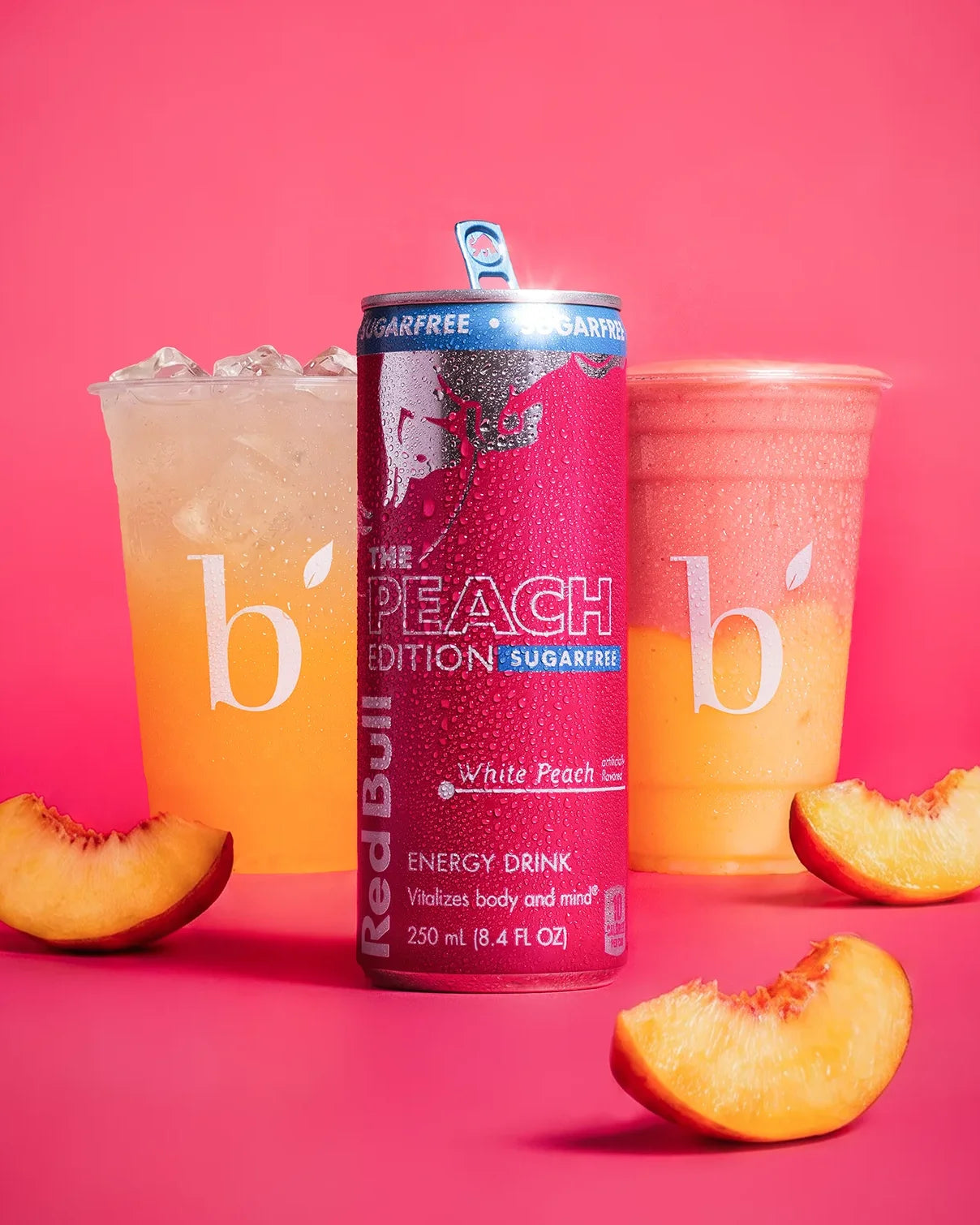 a can of peach red bull placed on a pink background in front of a smoothie and a refresher drink