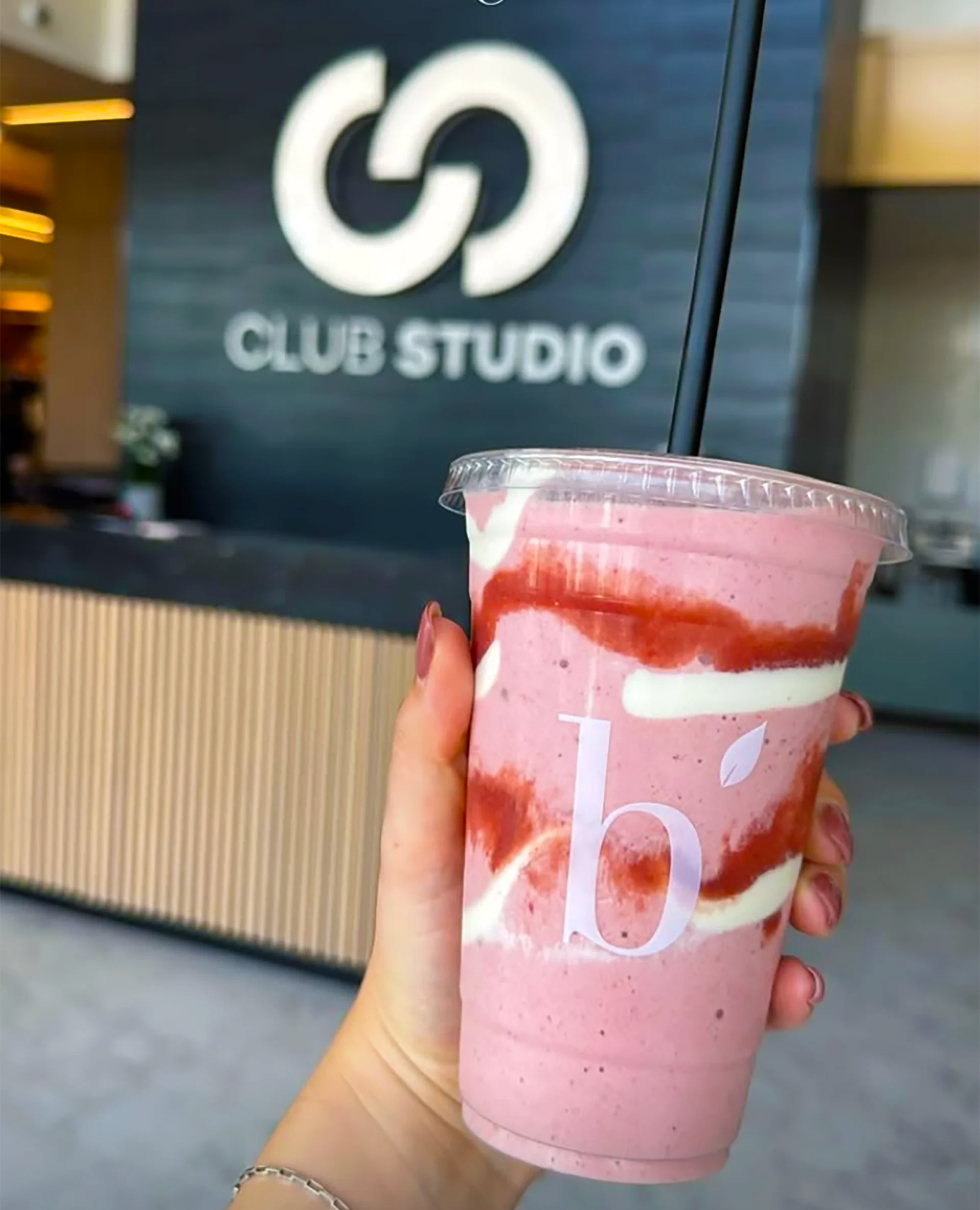 A hand holds a pink smoothie in a clear plastic cup with a black straw, featuring white cream swirls and red syrup drizzle. The background shows a blurred 'Club Studio' sign.
