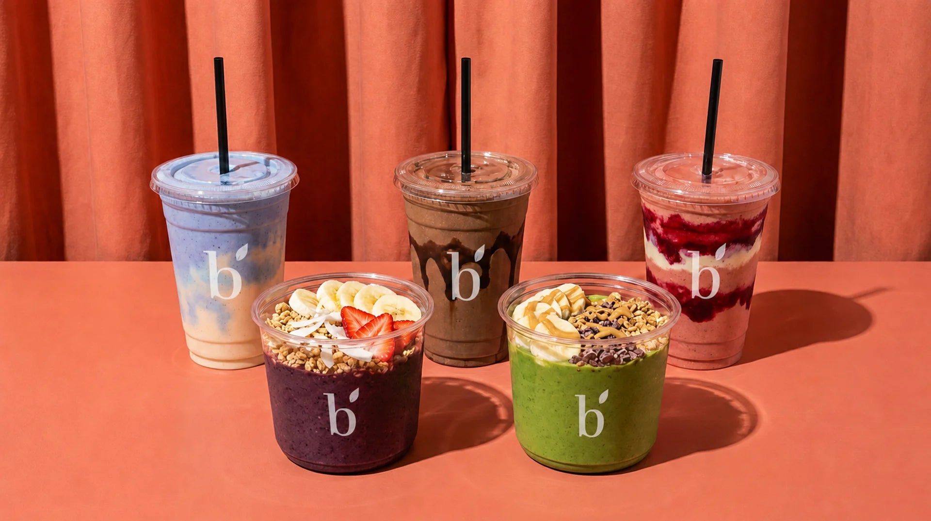 Five colorful smoothies and acai bowls with black straws are arranged on a peach table in front of peach curtains. The drinks feature different flavors and toppings, including banana, strawberries, granola, and green powder, and each has a white lowercase 'b' logo on the cup.