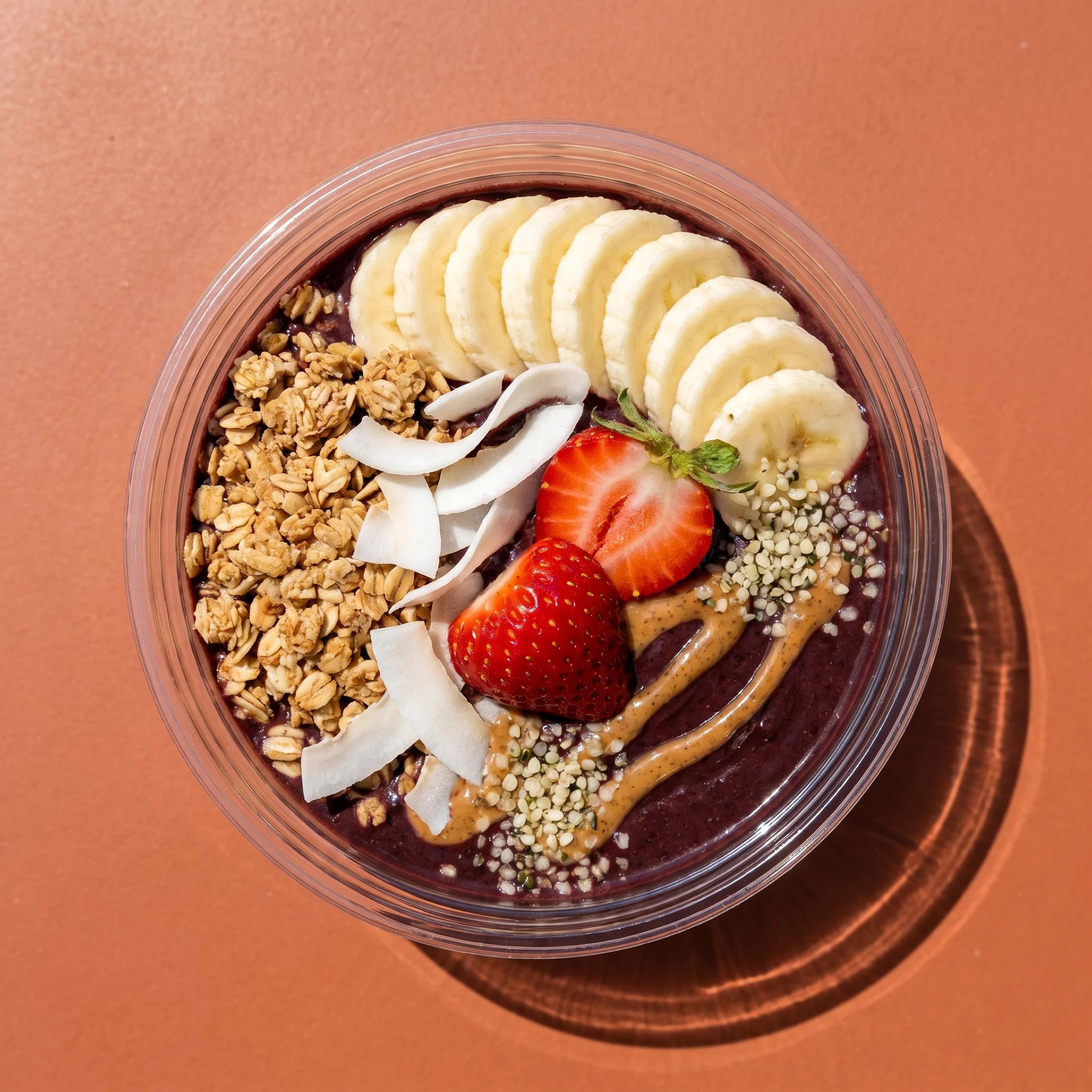 A clear plastic bowl filled with a purple acai smoothie base topped with sliced bananas, a strawberry half, coconut flakes, granola, hemp seeds, and peanut butter drizzle, photographed from above on a warm terracotta background.