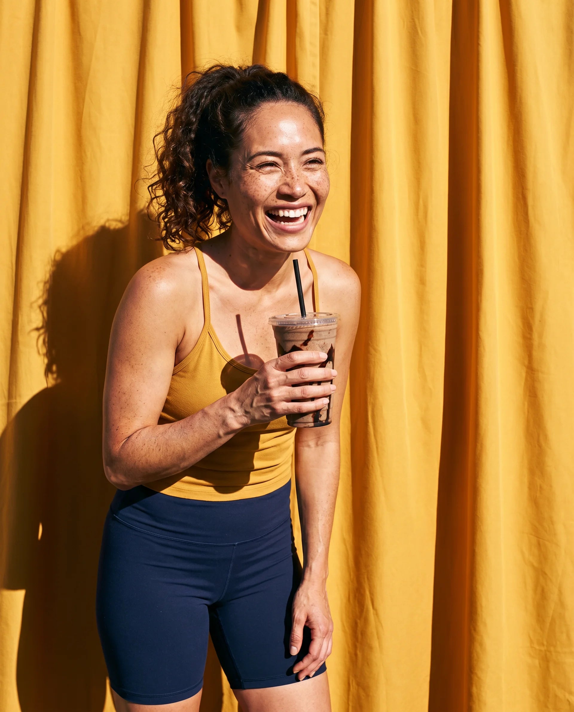 Woman in athletic wear holding a drink with a straw against a yellow curtain