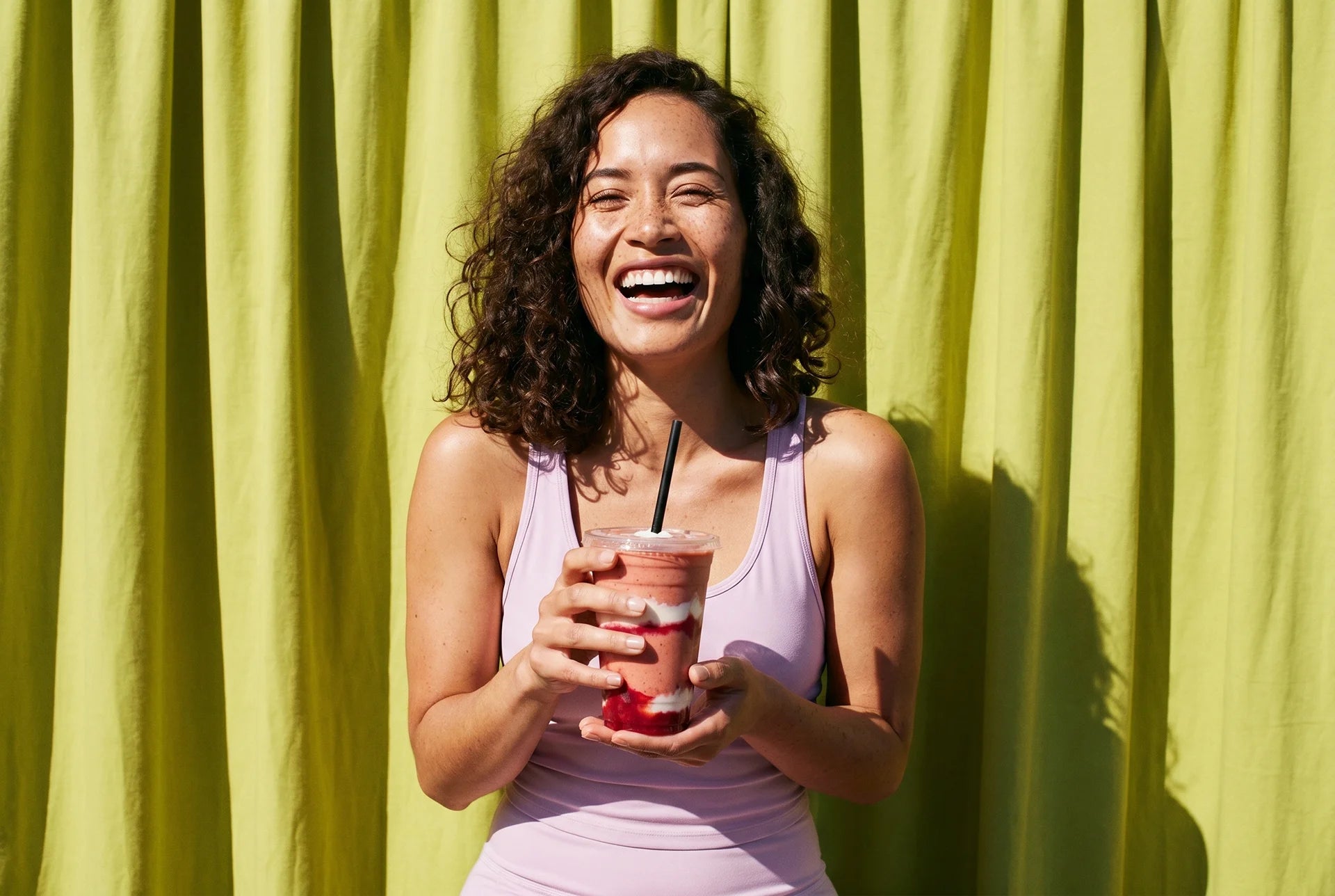 Woman in a purple tank top holding a red cup with a straw against a green curtain.