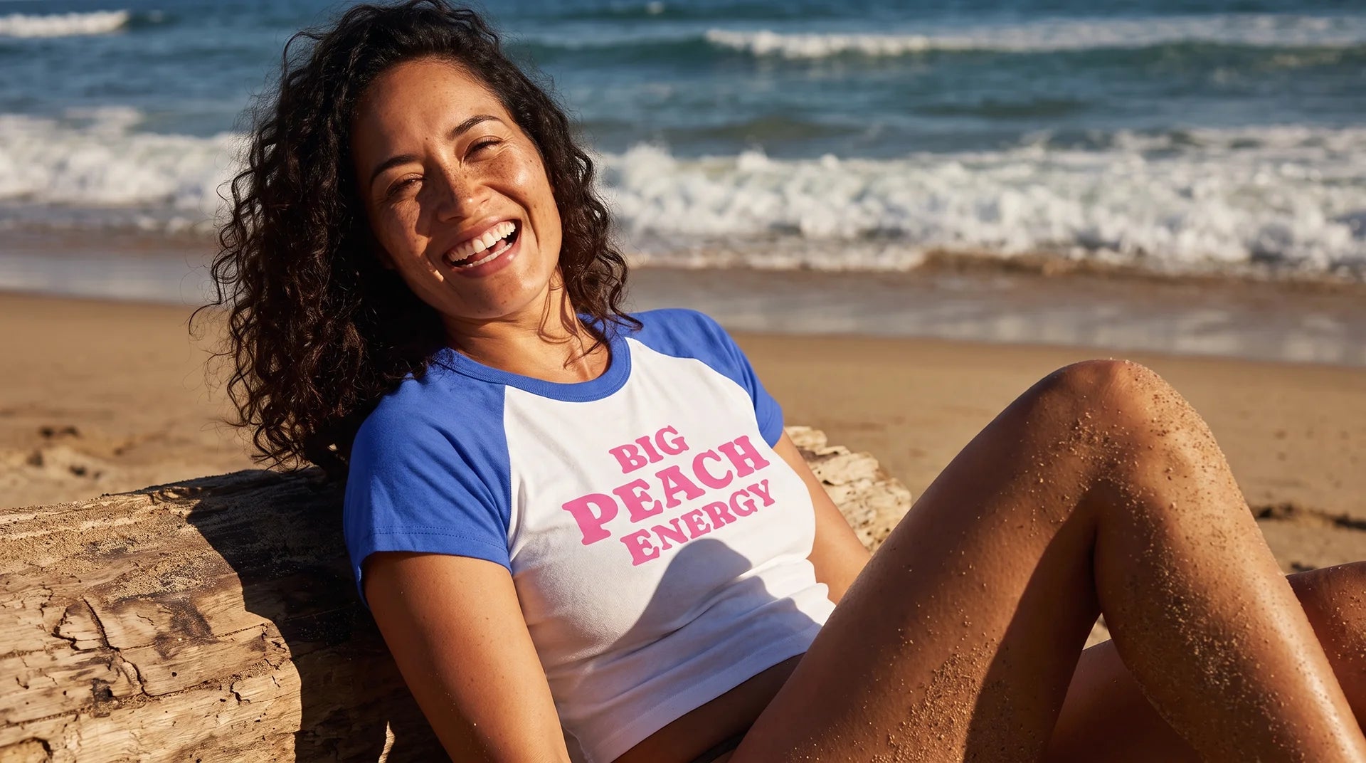 A woman smiling while sitting on a log on a sandy beach, wearing a raglan-style t-shirt with blue sleeves and a white body that reads 'BIG PEACH ENERGY' in pink text.
